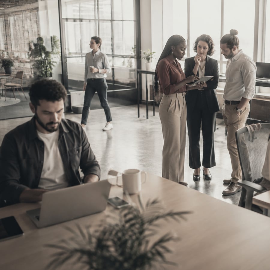 Man at desk, office setting, people talking in background. 